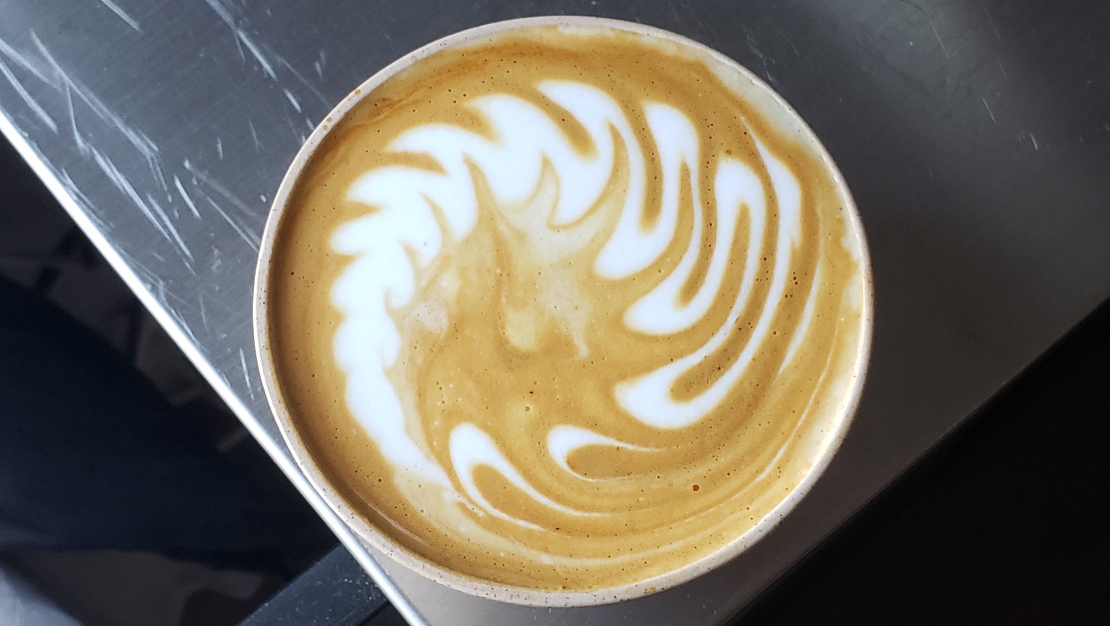 A swirling design of milk foam rests on top of a soft bed of coffee crema. It resembles a whirlpool or as if a fern leaf was twisted into a spiral.