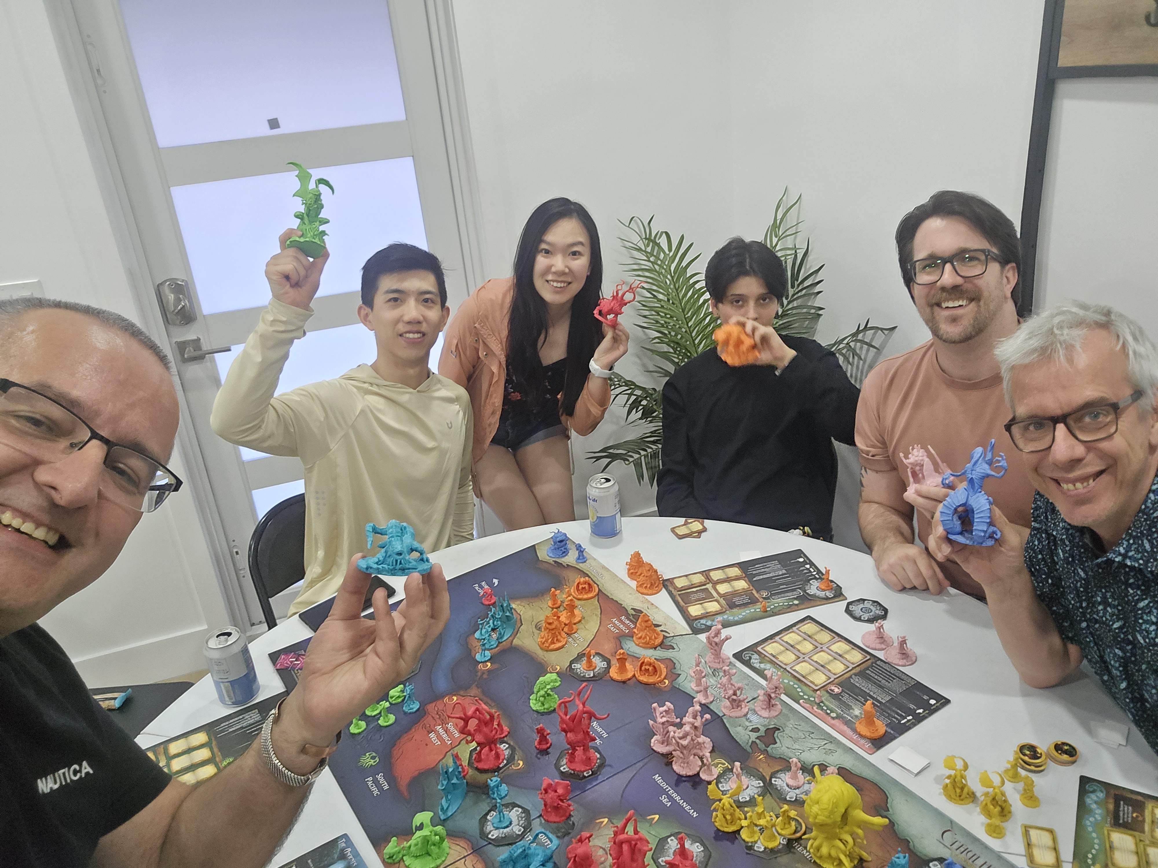 A group of friends, smiling with a large game board between them full of colorful plastic pieces shaped like horrible monsters.