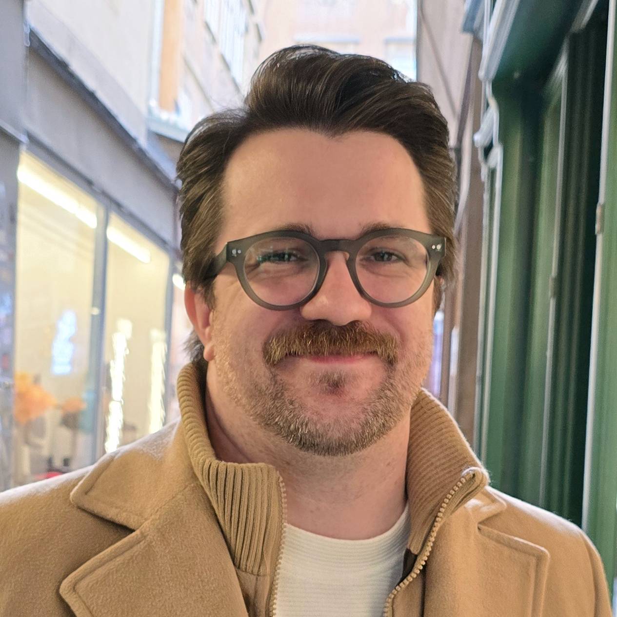 It's me! A white man smiling in a sunlit alleyway with medium-short brown hair, seaglass green trimmed glasses, and a tan overcoat.