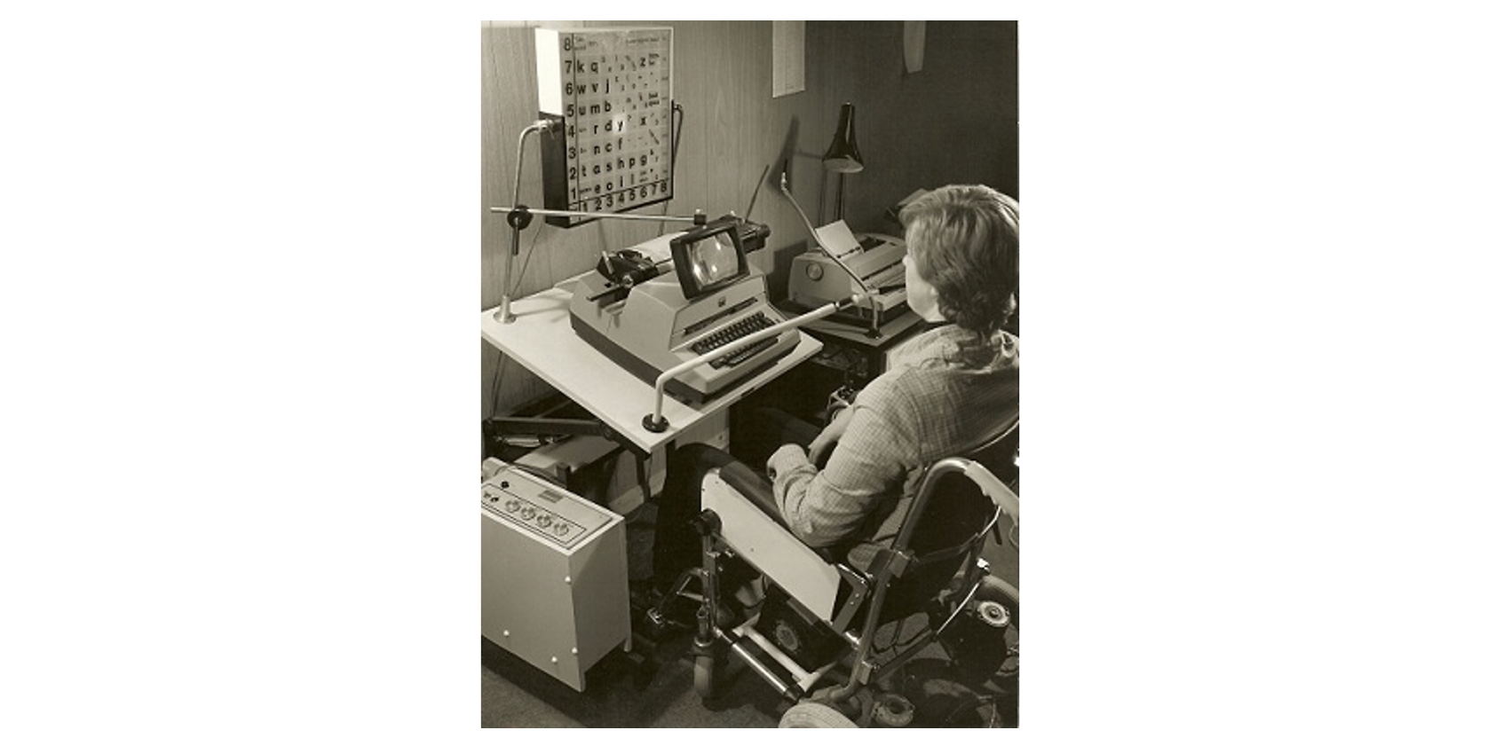 Photo of a person in a wheelchair operating an old computer using a desk-mounted sip and puff device called the POSSUM.